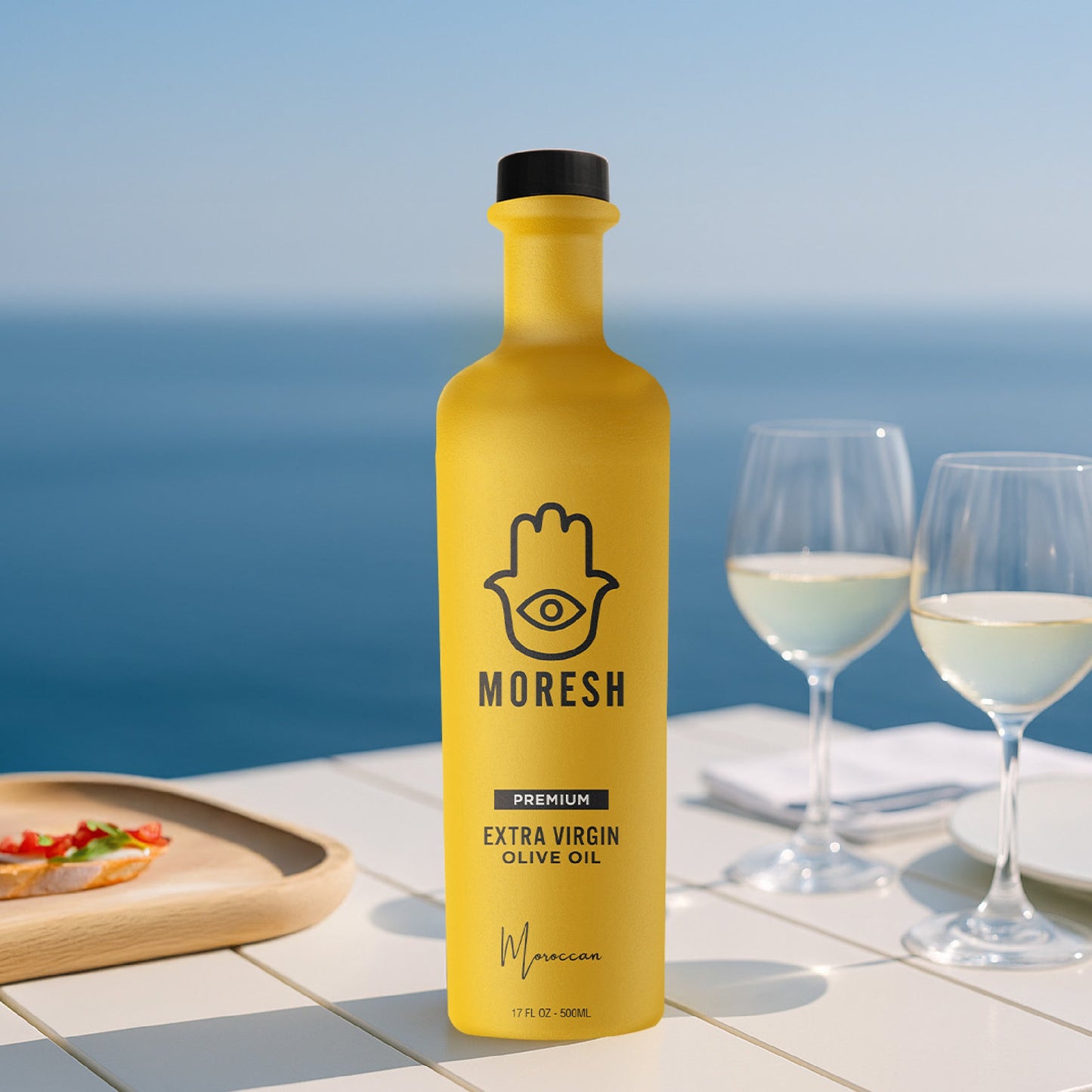 
                  
                    Yellow bottle of Moresh olive oil with two glasses of white wine and a plate of food on a table by the ocean.
                  
                