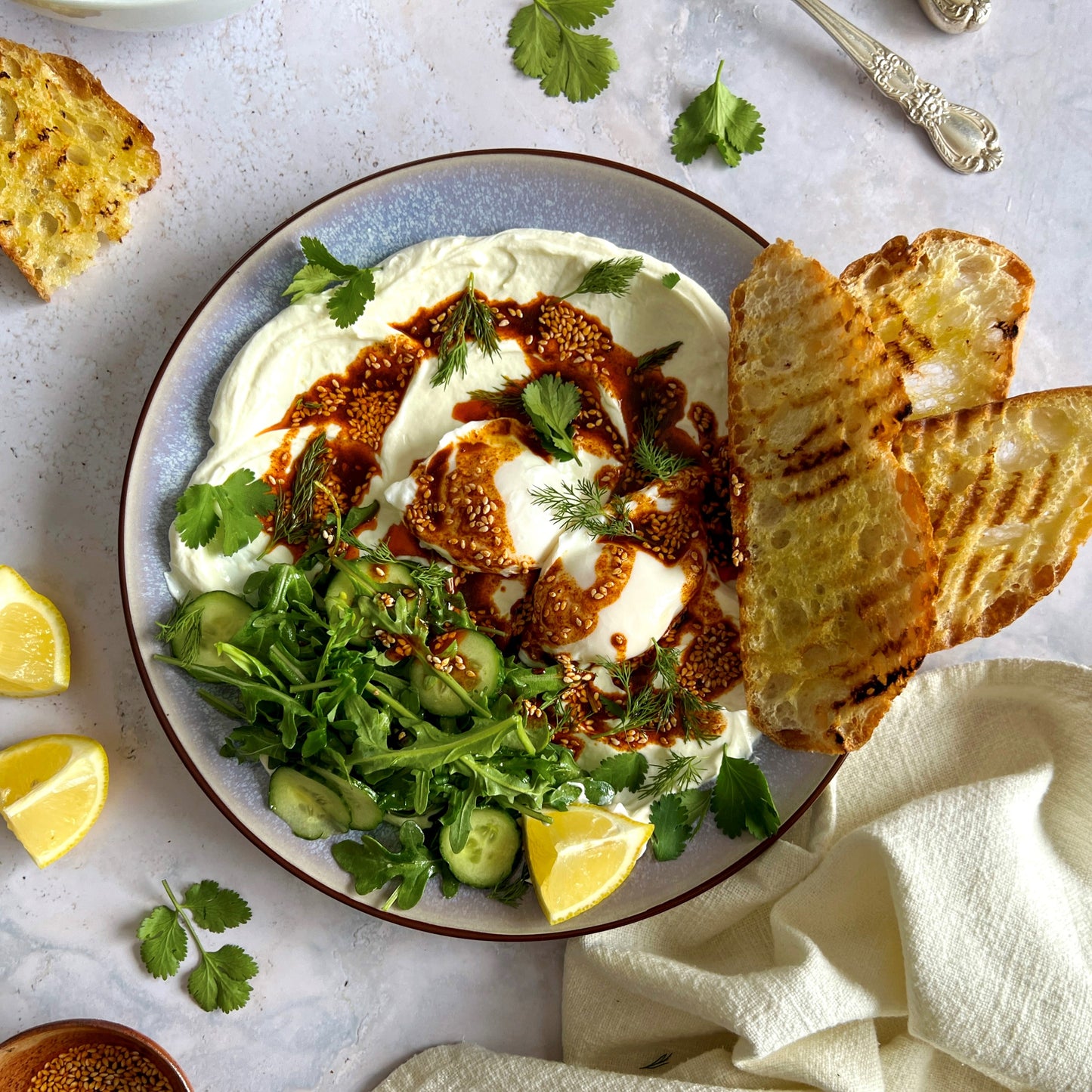 Delicious and savory Turkish eggs recipe featuring rich, velvety poached eggs nestled on top of yogurt and garlic-infused Terra Delyssa Extra Virgin Olive Oil, garnished with a drizzle of chili olive oil and fresh herbs and a side of warm, crusty bread for dipping – a nutritious and inviting Mediterranean culinary experience.